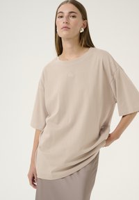 Beige ylisuurta t-paitaa, joka on valmistettu pehmeästä kankaasta. Siinä on lyhyet hihat ja pyöreä pääntie. Etupuolella on hienovarainen logodetail.