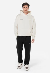 Sudadera crema con un bolsillo en el frente y pequeños diseños en texto. Combinada con joggers negros, terminados con zapatillas blancas y marrones.
