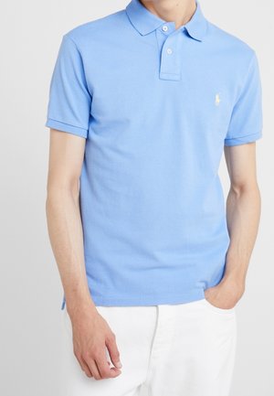 Mann trägt ein hellblaues Polohemd mit kurzen Ärmeln und weiße Hose, eine Hand in der Tasche, einfarbiger Hintergrund.