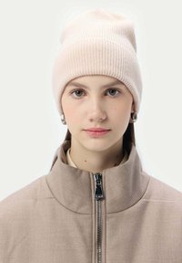 Ljusrosa stickad mössa med ribbad textur, bärs med en högkragad beige jacka med en silverdragkedja.