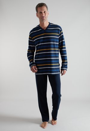 Mann steht barfuß, trägt ein dunkelblau gestreiftes Langarmhemd und einfarbige dunkelblaue Hose vor einem einfarbigen hellen Hintergrund.