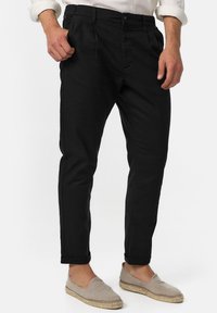 Man in zwarte tapere broek met opgerolde zomen, beige instap espadrilles en een witte shirt met opgerolde mouwen, staand op een witte achtergrond.