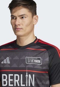 Maillot de football noir et rouge avec un motif géométrique, texte "BERLIN", un logo sur la poitrine et une marque Adidas sur l'épaule.