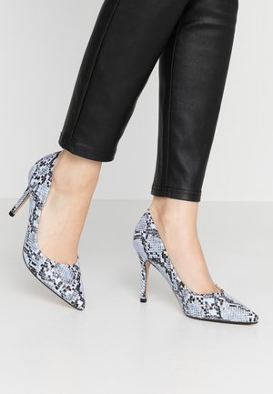 Spidse pumps i sort og hvid slangeskindsmønster med stilethæl og glat tekstur, båret med sorte tætsiddende bukser.