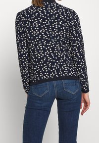 Kvinna i en mörkblå blomsterprydd långärmad tröja och blå denimjeans, stående med ryggen mot kameran.
