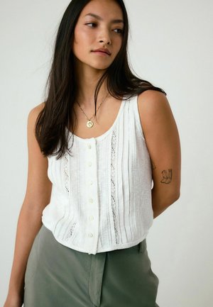 Junge Frau mit langem, dunklem Haar trägt eine weiße, knöpfbare ärmellose Bluse, grüne Hosen, eine goldene Anhängerkette und hat ein Tattoo am linken Arm.