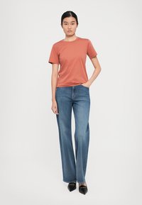 Ung kvinna står med en hand i fickan, iklädd en rostfärgad kortärmad skjorta, blå vida jeans och svarta spetsiga skor.