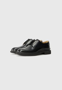 Pantofi negri din piele îmblănită, cu șireturi, cu un finisaj lucios, vârf rotunjit și o talpă din cauciuc negru, joasă și rezistentă. Design simplu, cu hardware minim.