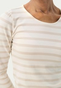 Person in einem beigen und weißen, horizontal gestreiften Langarmshirt mit rundem Halsausschnitt, von der Brust bis zum Hals gezeigt.