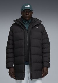 Sort polstret jakke med lynlåslukning, der har hætte og elastiske manchetter. Båret over en teal lynlåstop, med en mørk kasket, der viser Puma-logoet.