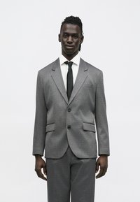 Hall textuuriga ülikond mustriga disainiga, ühe rinnaga jakk, sälgukraed ja must lips valge särgi kohal. Kaks esitaskut.