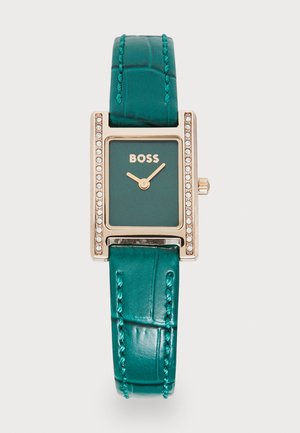 Montre avec un bracelet en cuir texturé vert, un boîtier rectangulaire en or rose, un cadran vert et des accents en cristal sur la lunette. Logo de la marque affiché.