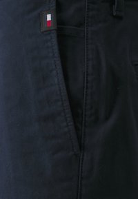 Pantaloni in tessuto blu navy con una texture liscia, dotati di una tasca laterale e un piccolo logo a patch in rosso, bianco e blu in vita.