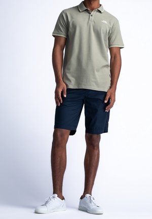 Homme portant un polo vert olive, un short bleu marine et des baskets blanches, debout sur un fond blanc uni.