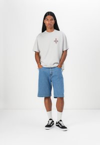 Lys grå t-skjorte med kort erme og en liten fargerik grafikk, kombinert med denimshorts. Modellen har på seg svarte sneakers og hvite ankelsokker.