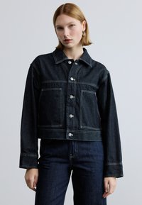 Fekete farmerdzseki, rövid fazonú, nagy első zsebekkel és jól látható fehér varrással, gombos záródással és szögletes gallérral.