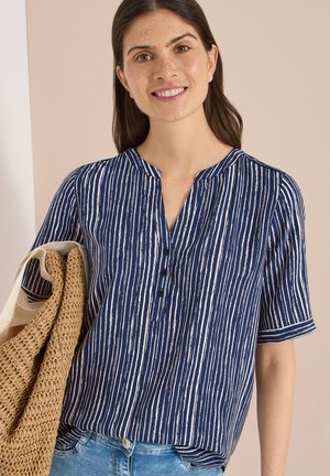 Uśmiechnięta kobieta w niebiesko-białej koszuli w pionowe paski i jasnych dżinsach trzyma dużą plecioną torbę na ramieniu.