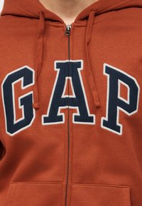 Άτομο που φοράει φούτερ με φερμουάρ σε χρώμα σκουριάς, με λογότυπο "GAP" σε ναυτικό και άσπρο χρώμα στο κέντρο του στήθους και κορδόνια να κρέμονται.
