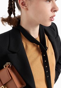 Blazer nero sopra un maglione senape, con una cinghia in tessuto regolabile e una borsa marrone con un accento di catena dorata.