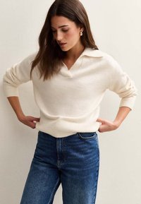 Sweter dziany w kolorze kremowym z dekoltem w serek, z kołnierzykiem. W połączeniu z niebieskimi dżinsami o wysokim stanie. Miękka faktura i luźny krój.