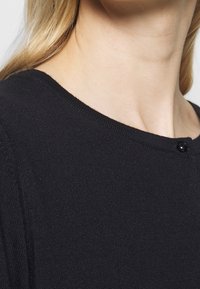 Schwarzer Strickpullover mit rundem Ausschnitt, sichtbarer Textur und einem kleinen schwarzen Knopfdetail am Ausschnitt.