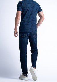 Marinblå t-shirt med ett subtilt blommigt mönster, kombinerad med mörkblå jeans och vita sneakers, sedd bakifrån.