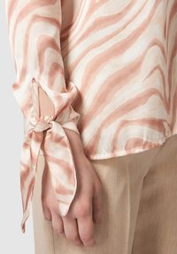 Blusa leggera color crema con righe ondulate rosa. Presenta un dettaglio ai polsini legato e una vestibilità rilassata, abbinata a pantaloni in lino beige.