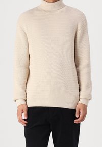 Pull à col roulé beige, texture côtelée, manches longues et poignets ajustés, associé à un pantalon noir. Design simple et moderne.