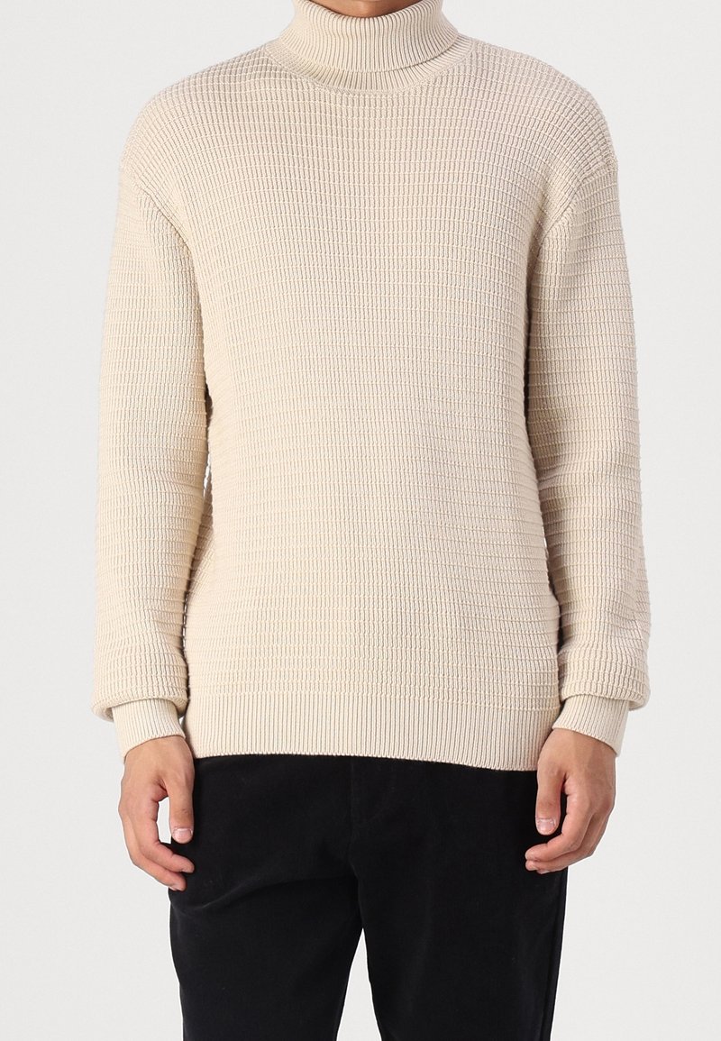 Pull à col roulé beige, texture côtelée, manches longues et poignets ajustés, associé à un pantalon noir. Design simple et moderne.