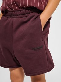 Person iført bordeauxfarvede sports-shorts med elastik i taljen og sidelomme samt "hummel"-logo på venstre ben, hånd hviler i lommen.