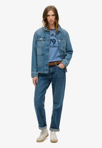 Jeansowa kurtka i spodnie z wysokim stanem w średnim odcieniu niebieskiego. Kurtka ma kieszenie na piersi. Model nosi niebieską koszulkę z grafiką i białe sneakersy.