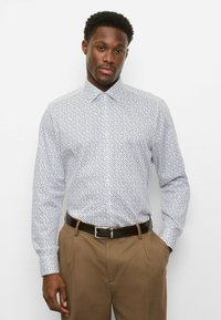 Langärmliges Hemd zum Knöpfen in Weiß mit einem blau-roten geometrischen Muster. Mit einem breiten Kragen und figurbetontem Schnitt. Kombiniert mit brauner Hose.