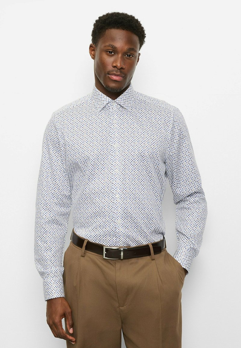 Langärmliges Hemd zum Knöpfen in Weiß mit einem blau-roten geometrischen Muster. Mit einem breiten Kragen und figurbetontem Schnitt. Kombiniert mit brauner Hose.