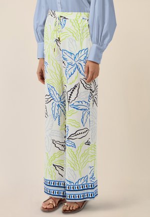 Frau trägt weiße, weit geschnittene Hosen mit blau-grünen und schwarzen tropischen Blattmustern, kombiniert mit einem hellblauen Langarmshirt und Sandalen.
