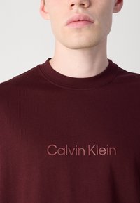 Burgundy cotton t-shirt with a ribbed neckline and embossed "Calvin Klein" logo in a lighter shade. Smooth texture, short sleeves.