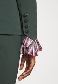 Zelený šitý rukáv s pěti černými knoflíky, který má na konci skládaný manžet v károvaném vzoru v červené, bílé a černé barvě. Viditelná ruka s ambrovou pletí.