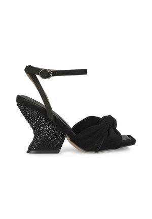 Sandalo a zeppa in suede nero con una parte anteriore annodata, cinghia sottile alla caviglia e tacco texturizzato e decorato con gioielli che presenta motivi geometrici.