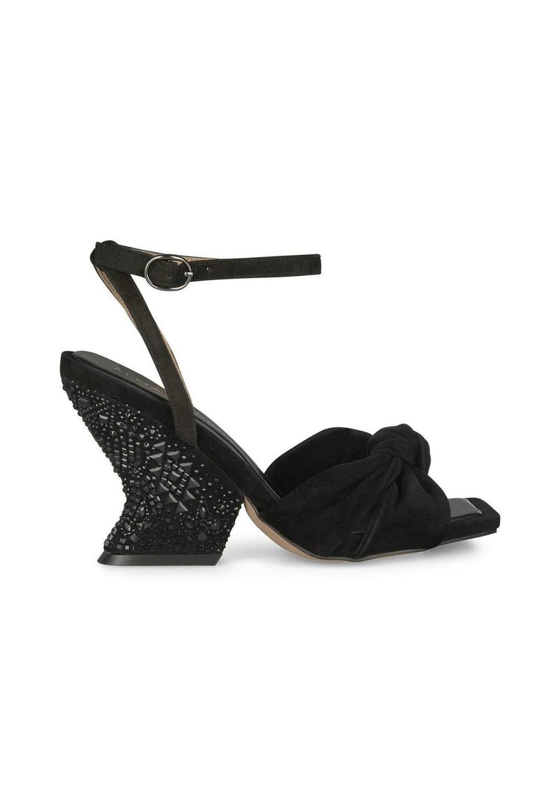 Sandalo a zeppa in suede nero con una parte anteriore annodata, cinghia sottile alla caviglia e tacco texturizzato e decorato con gioielli che presenta motivi geometrici.