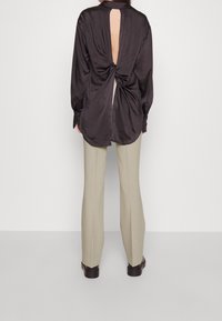 Zwarte satijnen blouse met een geknoopte achterkant en een open uitsnijding onder de kraag; gecombineerd met lichtbeige strakke broek en donkere schoenen.