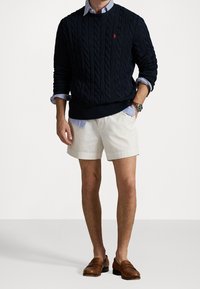 Pull en maille torsadée marine avec ourlet côtelé, porté sur une chemise rayée bleu clair, assorti à un short beige et des mocassins marron.