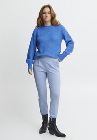 Blauer gestrickter Pullover mit strukturiertem Finish, kombiniert mit hellblauen, slim-fit Hosen. Schwarze Ankle Boots mit kleinem Absatz und Reißverschlussdetails.