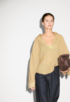Top beige leggero a maniche lunghe con scollatura a V e tessuto strutturato abbinato a jeans scuri. Una borsa a mano intrecciata marrone scuro è tenuta in una mano.