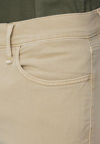 Beige bomullsbukser har en frontlomme med subtile sting og en glatt tekstur, kombinert med en mørkegrønn topp.