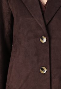 Blazer en daim marron avec un revers cranté, deux poches visibles et un bouton métallique rond proéminent à l'avant. Texture douce.