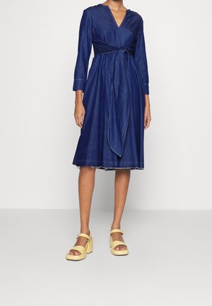 Femme portant une robe en jean bleu foncé longueur genou à manches longues, avec une ceinture nouée à la taille devant, associée à des sandales à talons hauts épaisses jaunes.