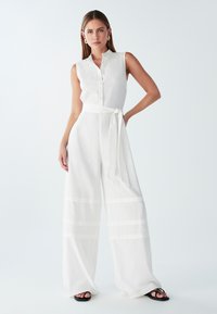 Mono de lino blanco con diseño sin mangas, frente de botones, piernas amplias y cintura ceñida con cinturón. Presenta rayas horizontales sutiles.