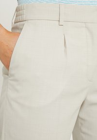 Lichtbeige op maat gemaakte broek met een gladde textuur, plooidetail aan de voorkant en een elastische tailleband voor comfort.