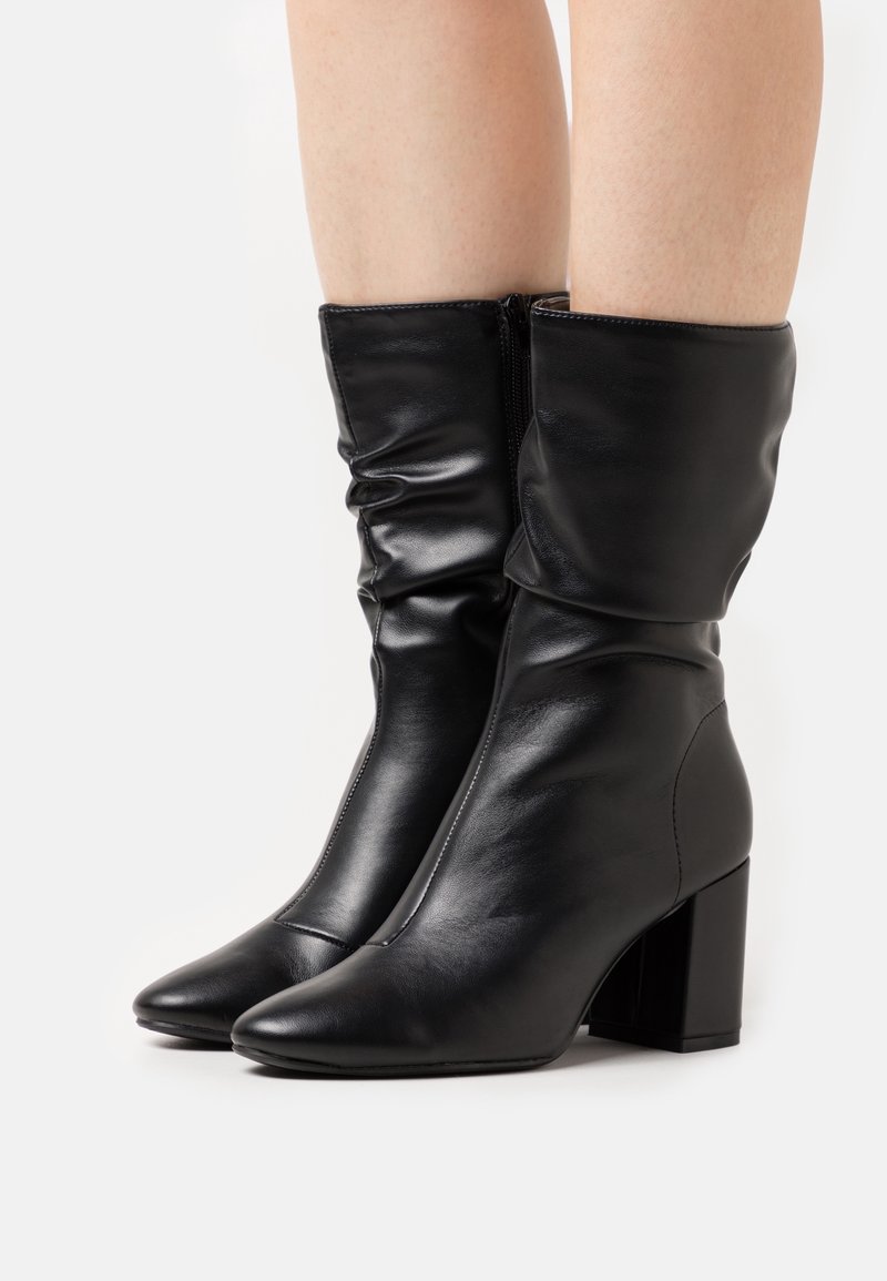 Bottes en cuir noir mi-mollet avec talons carrés et tiges légèrement souples portées sur jambes nues sur un fond uni.