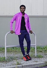 Paarse zip-up jas met zwarte accenten, zwart T-shirt, marineblauwe trainingsbroek en zwart-sneakers met rode accenten, staande naast een grijze reling.