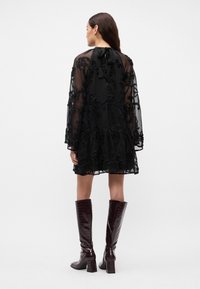 Crna prozirna haljina s cvjetnim vezom i dugim rukavima, koja ima naborani rub i izrez na leđima. Nosila se uz sjajne burgundske čizme do koljena.